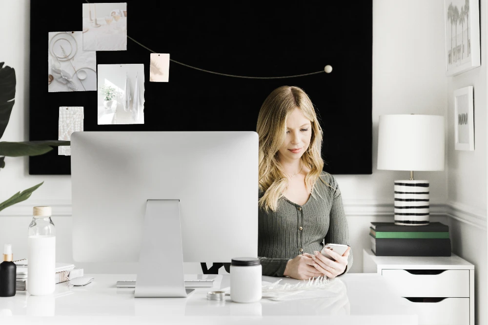 promotional image for email & website services provided by CSRA Marketing, a local service business website & amrketing agency. Image shows: a woman sitting at her computer desk & looking at her phone